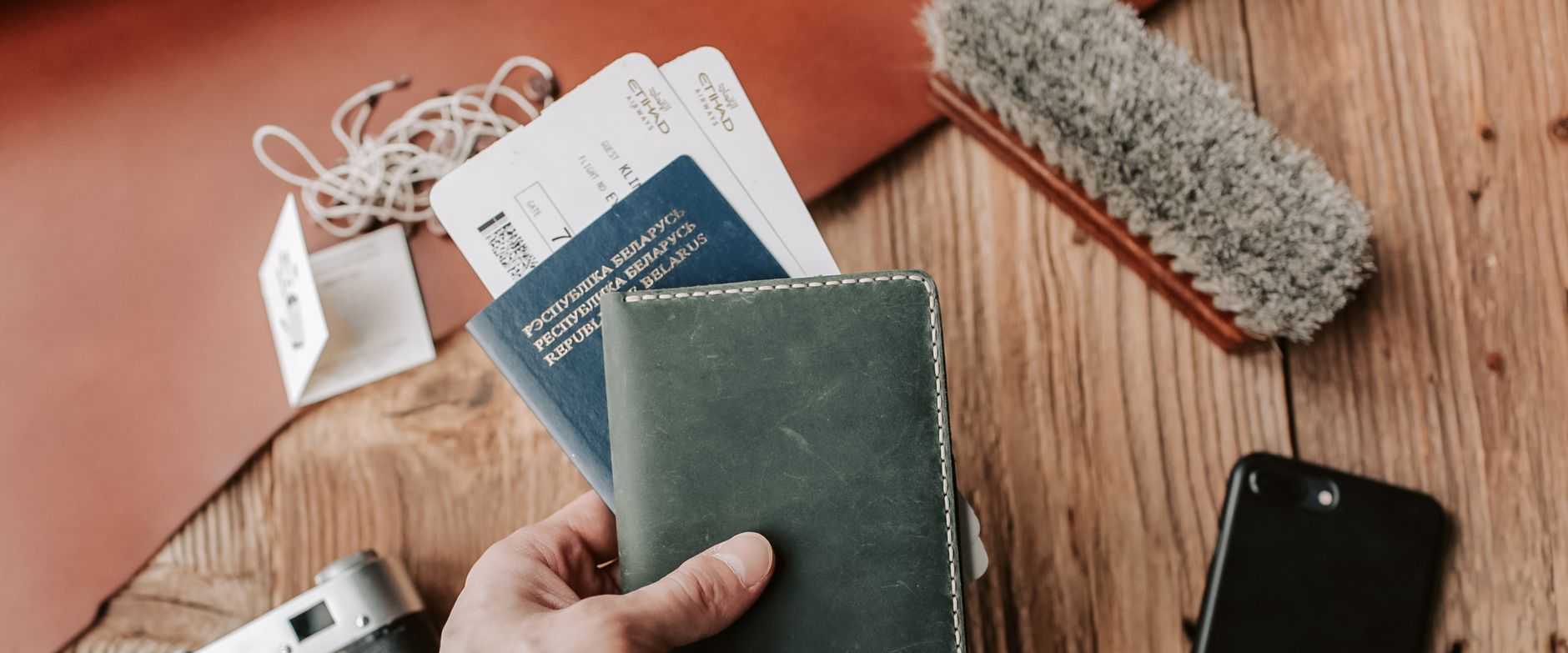 Nahaufnahme einer Hand, die einen Reisepass mit Reiseutensilien hält, darunter eine Kamera, ein Telefon und Tickets auf einem Holztisch.